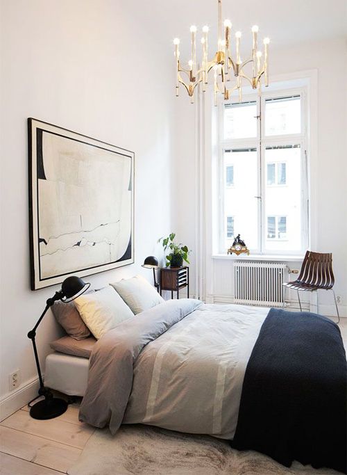 a small and elegant bedroom wiht a bed, monochromatic bedding, a statement artwork, a chandelier and a cool chair