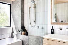 a small bathroom with skinny white tiles and grey stone ones, a bathtub, a floating vanity, a mirror and a window