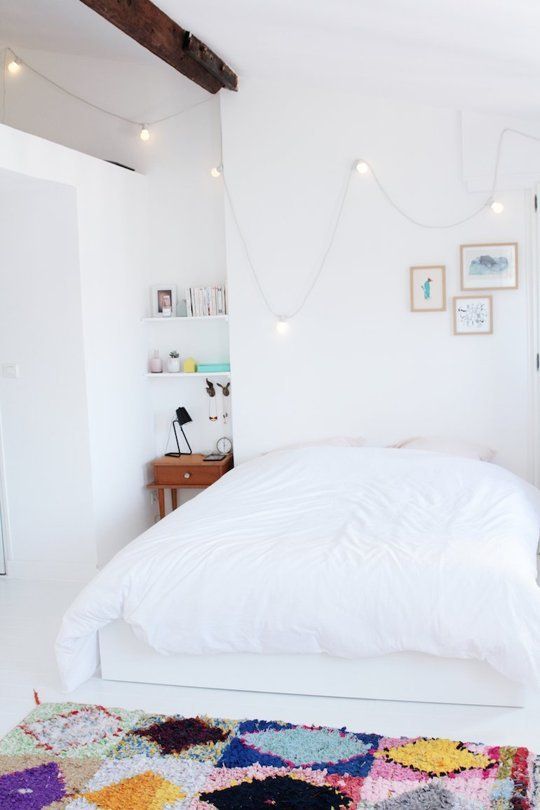 a small bedroom with a wooden beam, a bed with white bedding, a tiny nightstand in the corner, a bright rug and lights