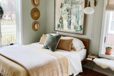 a small boho bedroom with light green walls, a bed with neutral bedding, a bench, an artwork and a gallery wall of hats