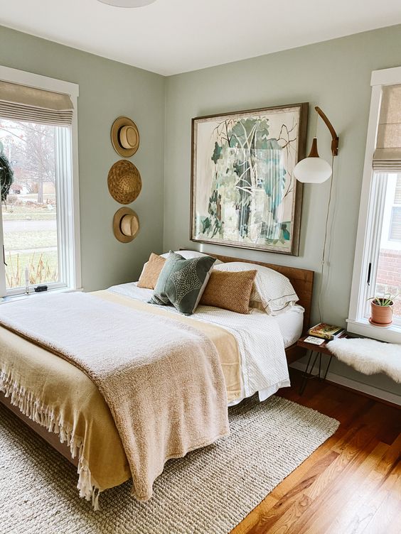a small boho bedroom with light green walls, a bed with neutral bedding, a bench, an artwork and a gallery wall of hats