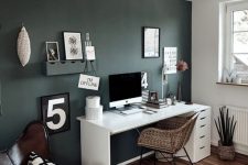 a small boho home office with a dark green accent wall, a white desk, a woven chair, a leather chair with a pillow