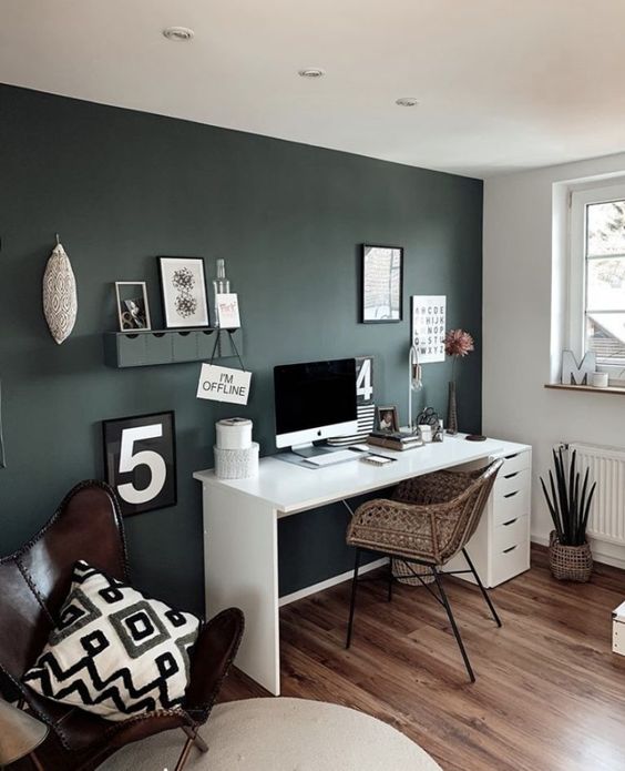 a small boho home office with a dark green accent wall, a white desk, a woven chair, a leather chair with a pillow