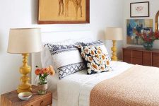a small bright bedroom with a bed with printed bedding, stained nightstands, table lamps, a dresser and bold art