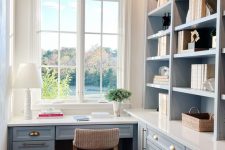 a small farmhouse home office with pastel blue cabinetry and a built-in desk, some lights and lamps and a lovely view from the window