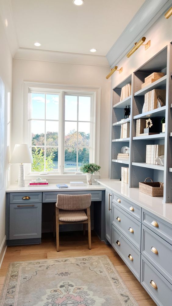 A small farmhouse home office with pastel blue cabinetry and a built in desk, some lights and lamps and a lovely view from the window