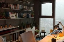 a small home office with a floor to ceiling window, open shelves, a storage unit, a desk, a chair and a table lamp