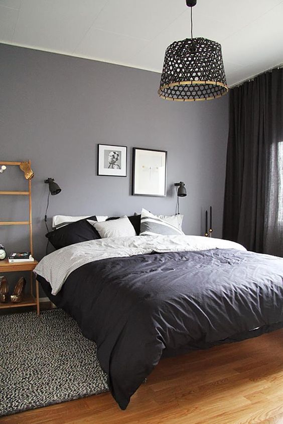 a small moody bedroom with a grey wall, a bed with monochromatic bedding, a woven pendant lamp and black curtains