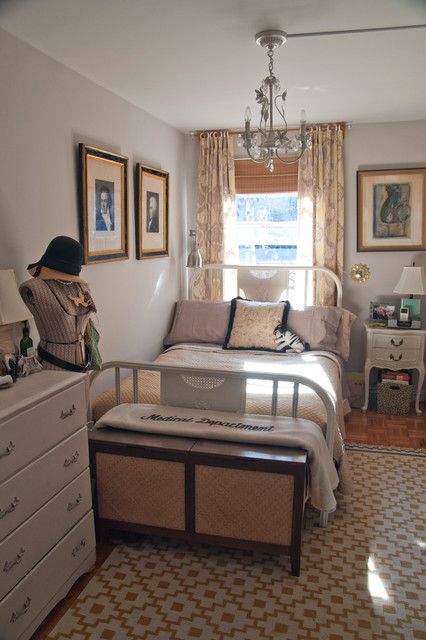 a small vintage bedroom with a forged bed with neutral bedding, a printed rug, a white dresser, a vintage nightstand and a chandelier