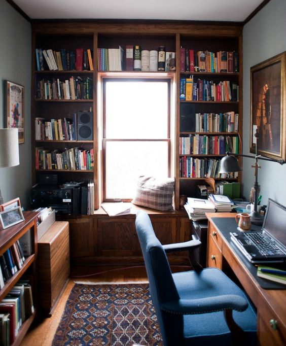 a small vintage home office with grey walls, a large bookcase framing the window, a stained desk, a blue chair and a windowsill seat