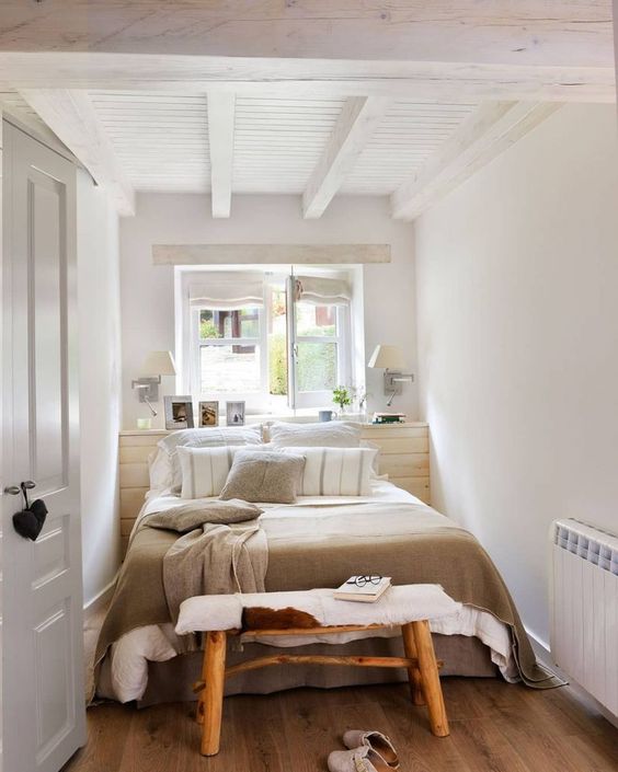 a tiny bedroom with a stained accent wall, a bed with neutral bedding, a bench, a couple of lamps and a window