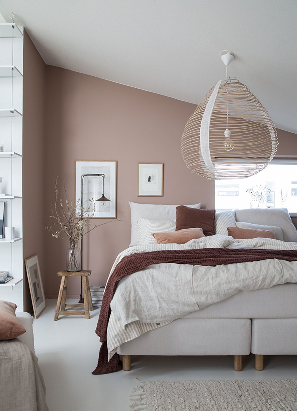 a warm and soft Scandinavian bedroom with mauve walls, a creamy bed, a woven lamp, artworks and a shelving unit by the window