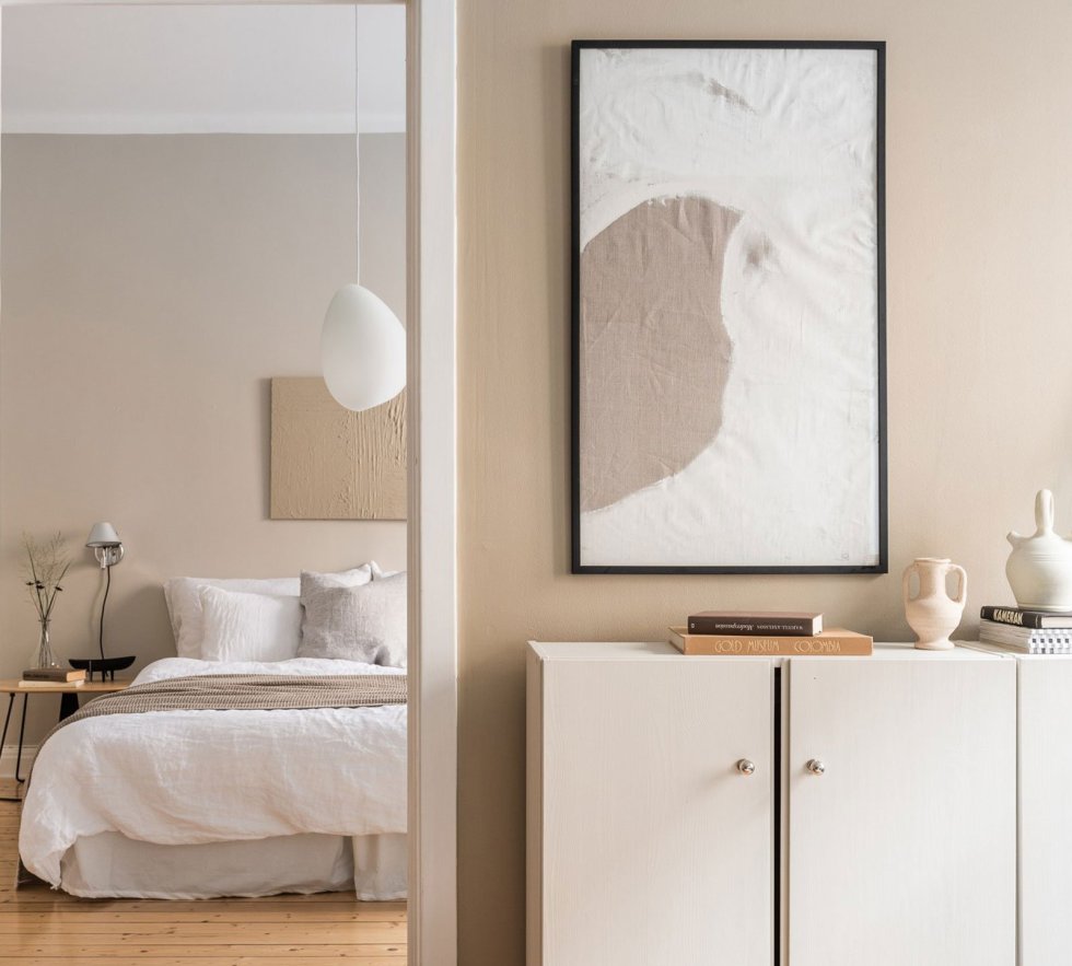 a warm beige bedroom with tan walls, a bed, wooden nightstands, a textural artwork and some dried blooms is welcoming