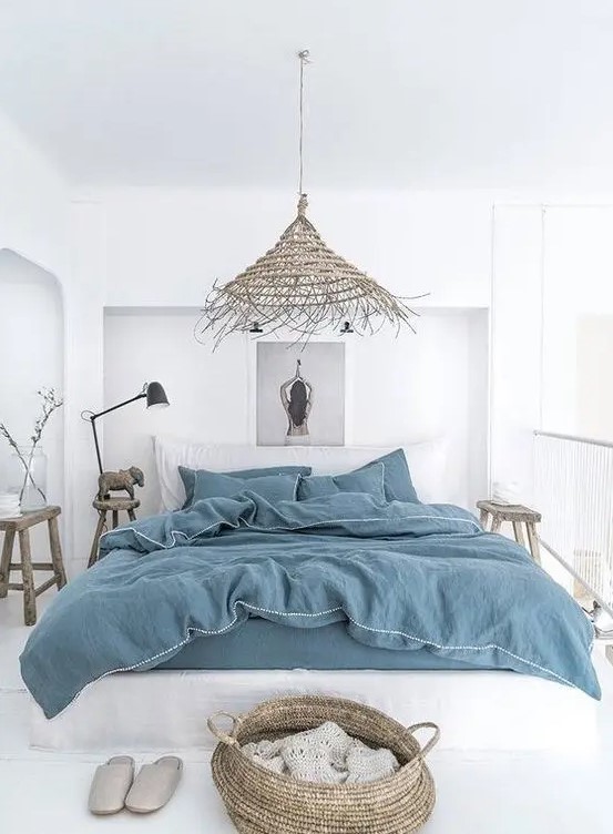 A white boho beach bedroom with all white everything, blue bedding, baskets, a wicker lamp and wooden furniture