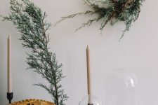 a wreath with dried branches and greenery, neutral candles, a grene branch and a pinecone in a cloche for a Nordic feel