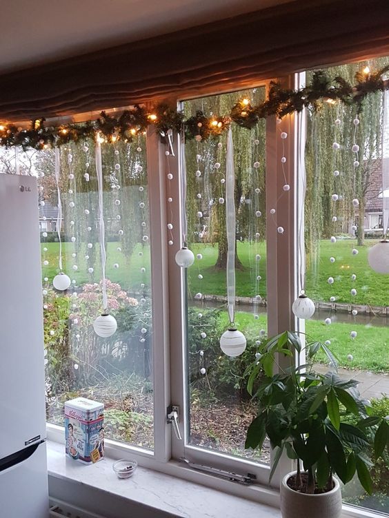 an evergreen garland with lights, pompoms and ornaments hanging down is a cool window decoration