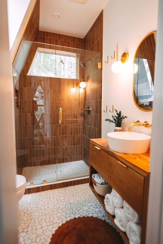 an eye-catching attic bathroom with a shower with rust tiles, a stained vanitu, an oval mirror and some gold fixtures