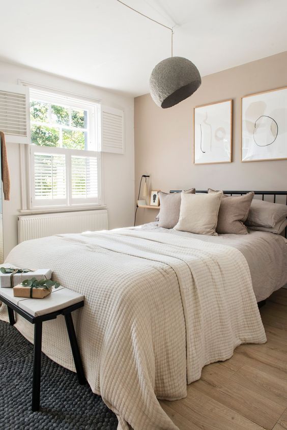 an inviting modern bedroom with a greige accent wall, a bed with greige bedding, a bench, a pendant lamp and some art