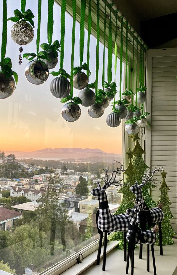 whimsical Christmas window styling with silver ornaments hanging down on green ribbons, green mini trees and deer on the windowsill