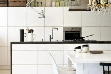 a Nordic off-white kitchen with a wooden ceiling, white sleek cabinets and a black countertop, a vintage chandelier and white chairs on wooden legs