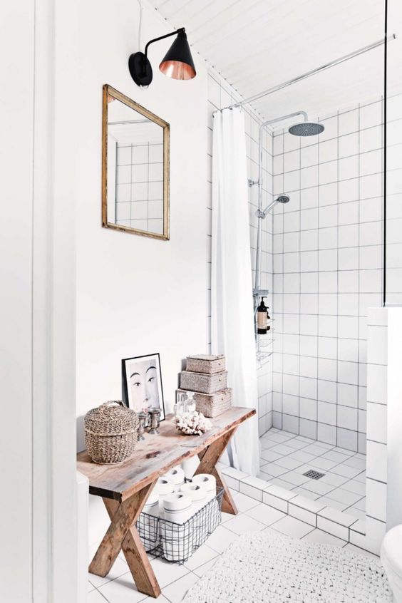 a Scandinavian bathroom done with white square tiles, a wooden bench with baskets, a black sconce, a mirror is lovely