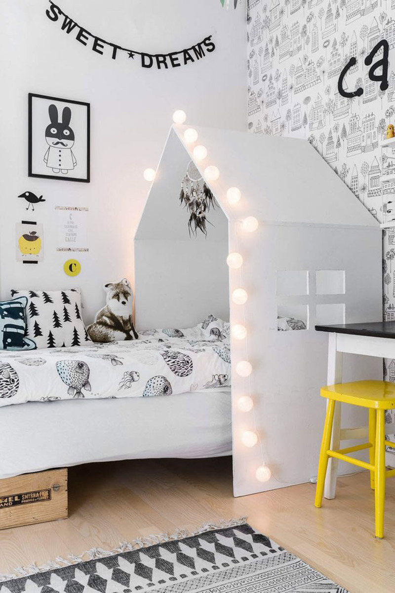 a Scandinavian kid's room with a bed with a house-shaped cover with lights, a desk, a yellow chair, a wall with printed wallpaper, some art