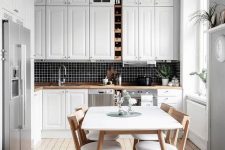 a Scandinavian kitchen with white shaker style cabinets, a black tile backsplash, a neutral dining set and a crystal chandelier