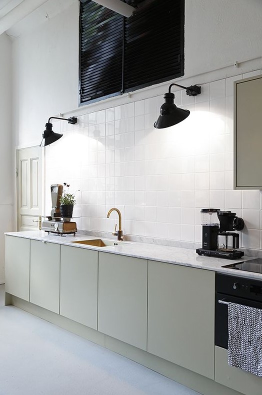 a Scandinavian kitchen with white walls and white square tiles, a row of olive green cabinets, black sconces
