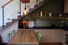 a catchy modern kitchen placed under the staircase, with a green tile backsplash, greige lower cabinets, black countertops and an open shelf