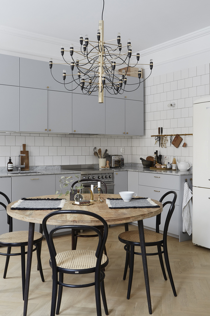 a chic Scandinavian kitchen with dove grey cabinetry, white square tiles, a very catchy chandelier and a retro wooden dining set