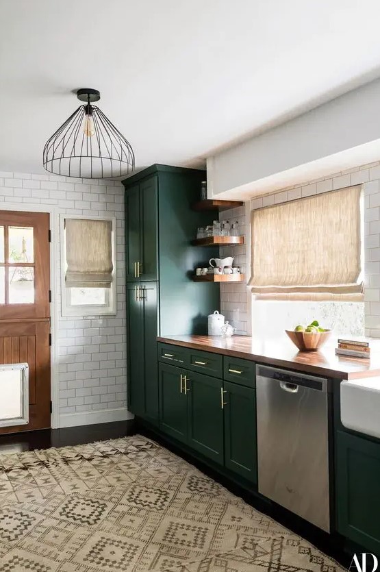 A chic dark green farmhouse kitchen with a buctcherblock countertop, woven shades and metal pendant lamps plus a boho rug.