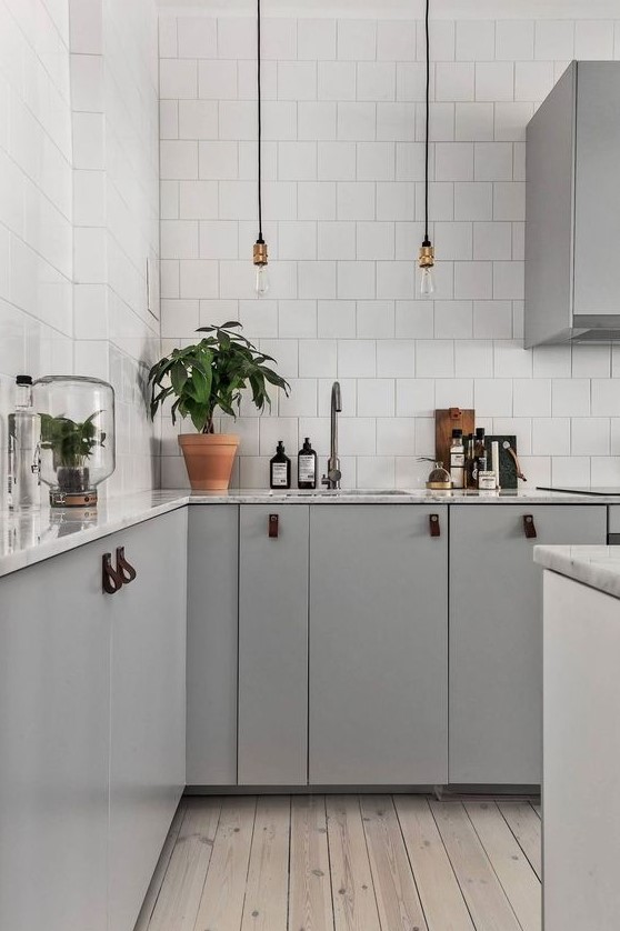 a contemporary Scandinavian kitchen with grey cabinets with leather pulls, bulbs, white tiles and a wooden floor