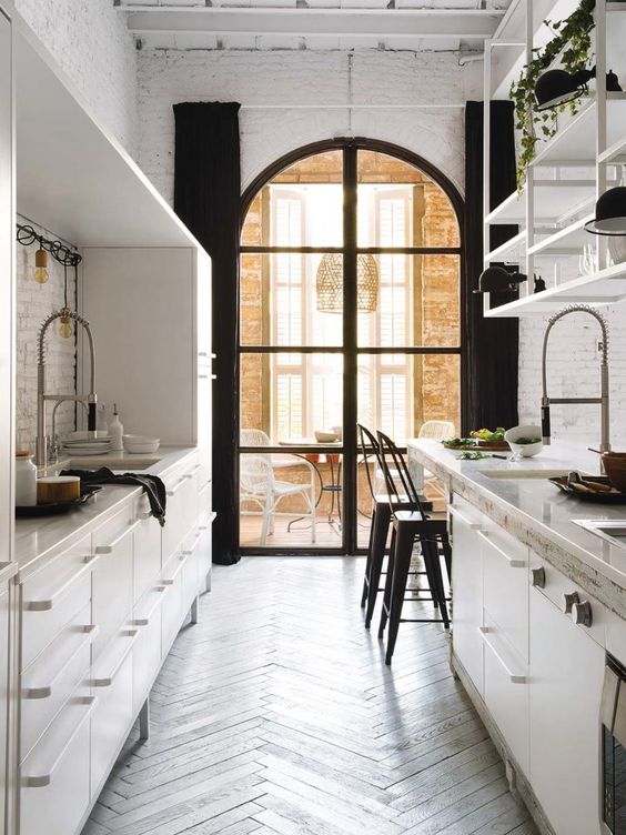 a contrastign galley kitchen with metal cabinets, an arched window, a dining zone by the window and greenery