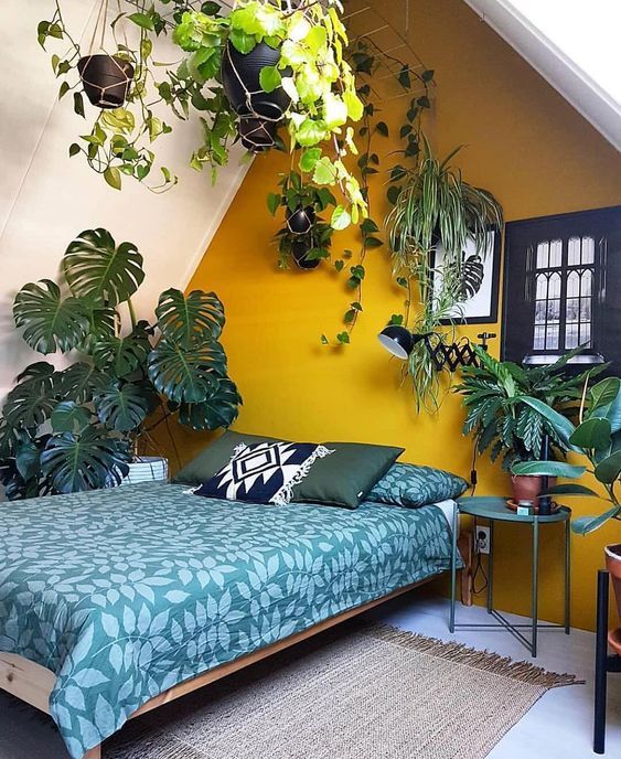 a cool boho attic bedroom with a mustard accent wall, some modern furniture, lots of potted plants and botanical bedding