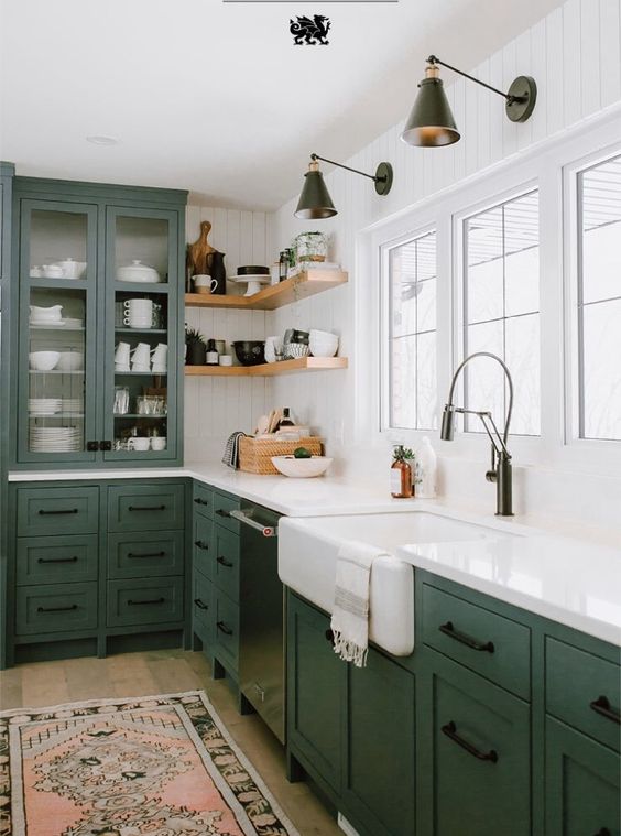 A green farmhouse L shaped kitchen with shaker cabinets, corner shelves, black sconces, white countertops and a backsplash