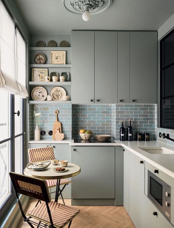 A grey L shaped kitchen with sleek cabinets, white stone countertops, a blue and grey subway tile backsplash, a tiny table and chairs