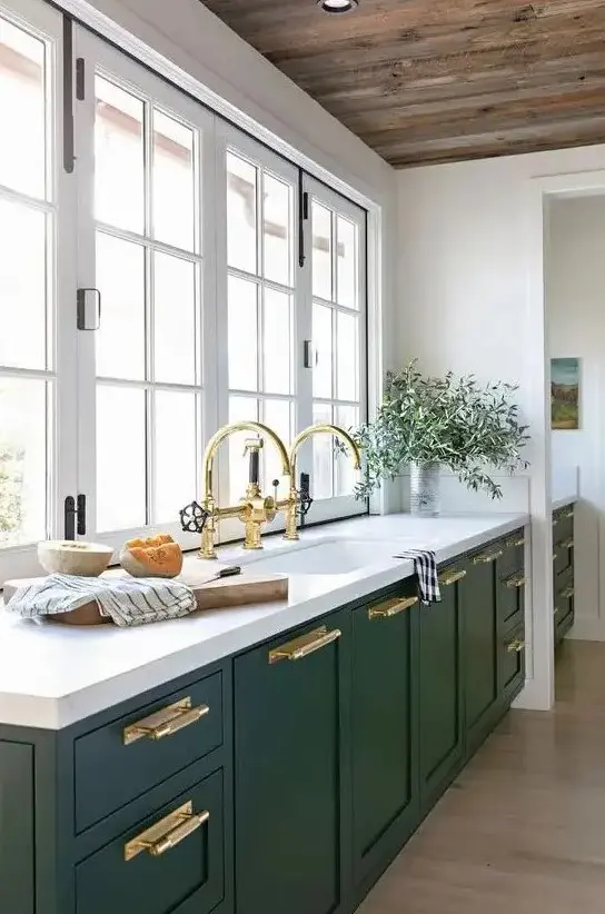 Hunter green cabinets and white stone countertops create a modern kitchen. Gold handles and fixtures provide a luxurious accent. A window replaces the backsplash, flooding the space with light.