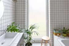 a light-filled bathroom with white square tiles, a tub clad with tiles, a vanity clad with them, too, some potted greenery