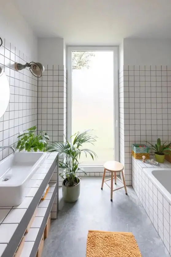 A light filled bathroom with white square tiles, a tub clad with tiles, a vanity clad with them, too, some potted greenery
