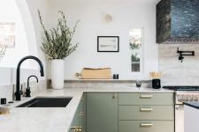 a light green L-shaped kitchen with neutral stone countertops, a black stone hood, black fixtures and some greenery