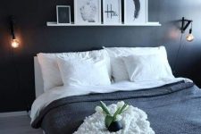 a minimal bedroom with a black accent wall, a ledge with artworks, a fluffy lamp and black and white bedding