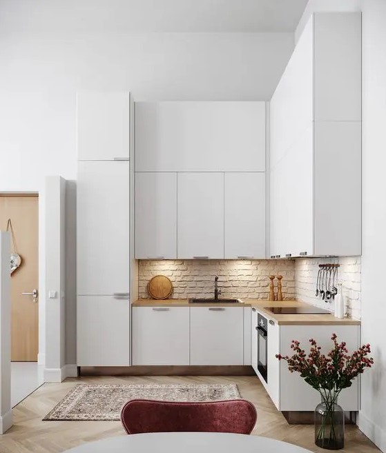 A minimalist white L shaped kitchen with a brick backsplash, butcherblock countertops and built in lights