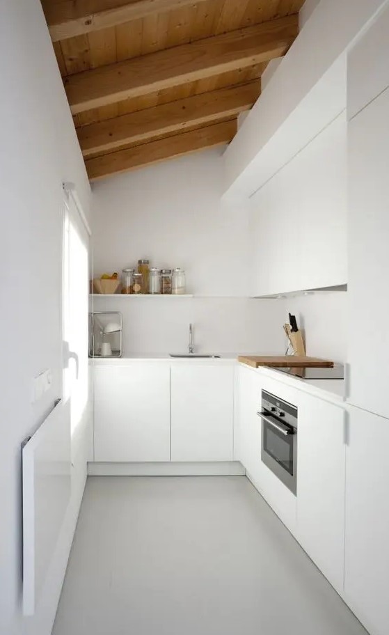 A minimalist white L shaped kitchen with a wooden ceiling and beams plus a window is very cool and doesn't look too small