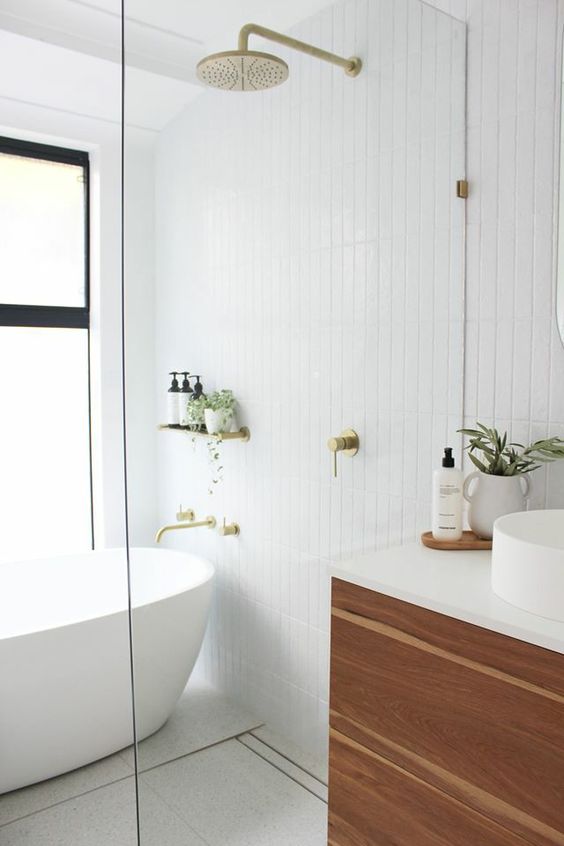 a modern Scandinavian bathroom clad with white tiles, an oval tub, a stained vanity, a white tub and sink, brass fixtures