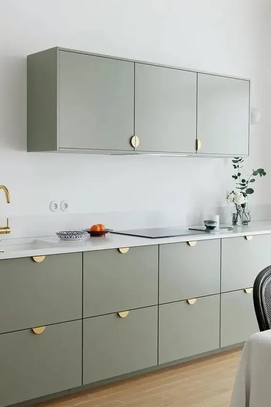 A modern kitchen features sage green cabinets with gold pulls for a sleek look. White stone countertops and a small backsplash add elegance. The minimalist design keeps the space chic and uncluttered.