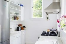 a narrow white kitchen with shaker style cabinets, white stone countertops, windows, bright blooms and a fridge