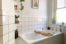 a neutral bathroom with a tub clad with green shiplap, white square tiles, a printed tile floor, potted plants and artwork