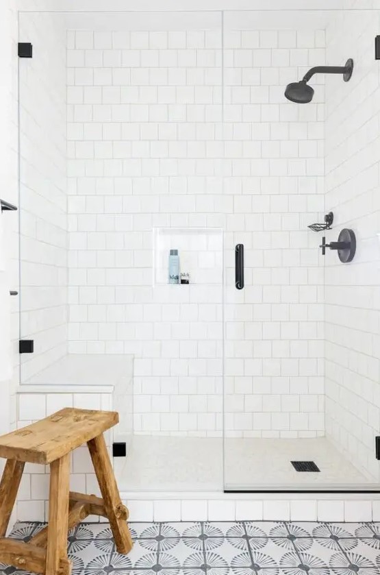 a neutral bathroom with a white square tile shower, a bold tile floor, black fixtures and a wooden stool welcomes in