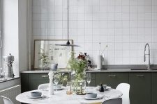 a pretty Nordic kitchen with dark green lower cabinets, white square tiles, a round table and white chairs and grey lamps