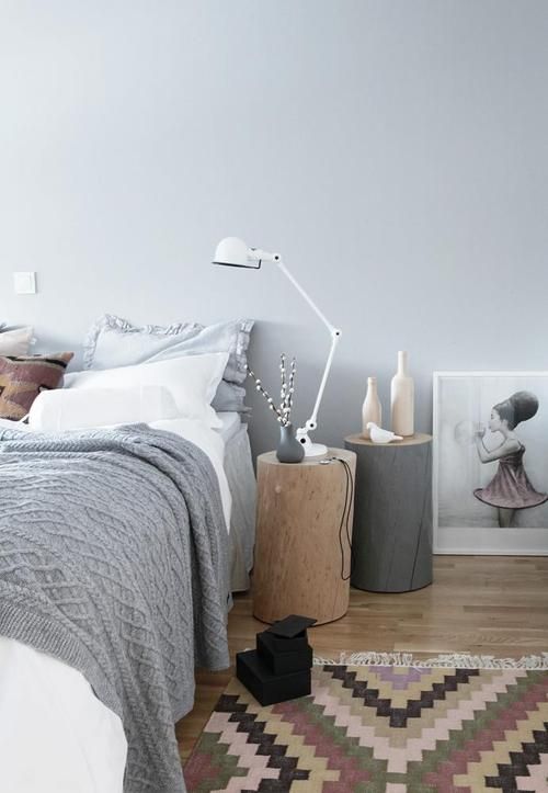 a relaxed Scandinavian bedroom with a bed styled with grey and white bedding, stumps with a lamp and bottles and a printed rug